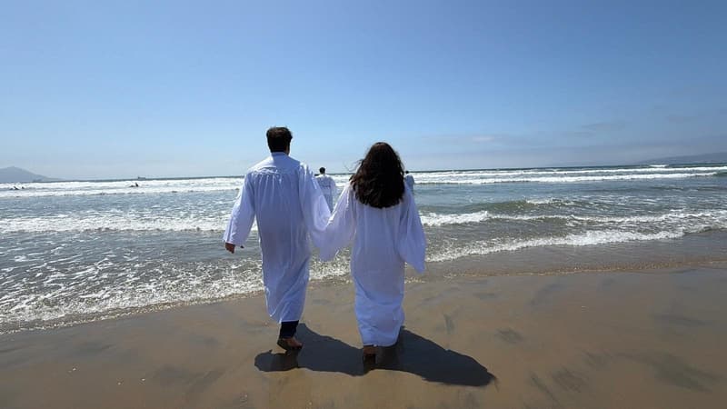 Dos personas con túnicas blancas caminando hacia el mar, vistas de espaldas.