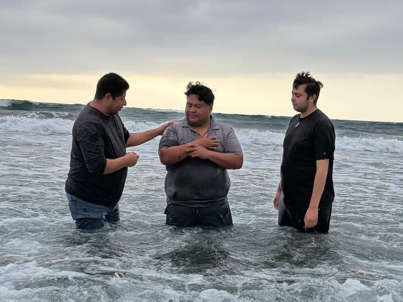 Un bautismo en el oleaje: un hombre con los brazos cruzados sobre el pecho, entre dos hombres, en las olas.
