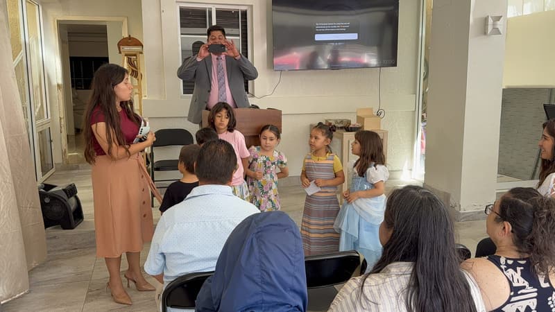 A teacher speaking with a group of children at the front of the church while the pastor takes a photo behind them.