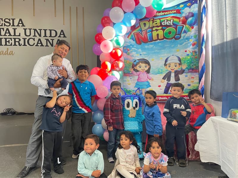 Niños y un hombre con un niño pequeño en brazos frente a un fondo del Día del Niño en la iglesia de Ensenada.