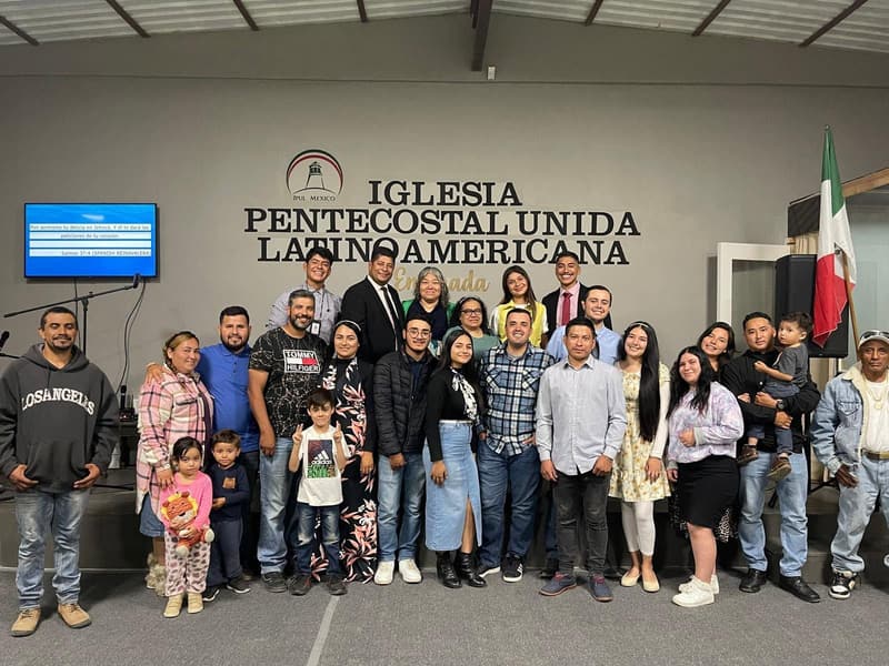 Miembros de la iglesia de Ensenada, adultos y niños, sonriendo juntos frente al nombre de la iglesia en la pared.