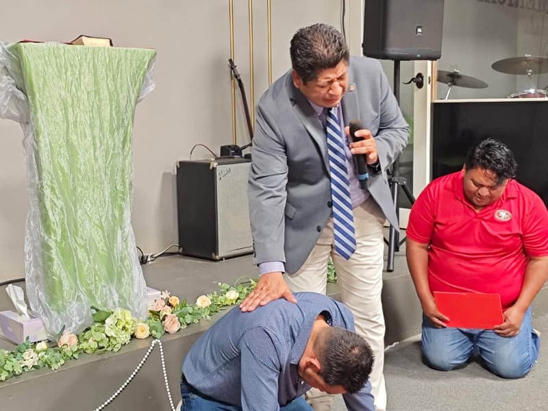 A man in a suit prays with his hand on the shoulder of a man kneeling at the front of the Ensenada church, while another man kneels and prays nearby.