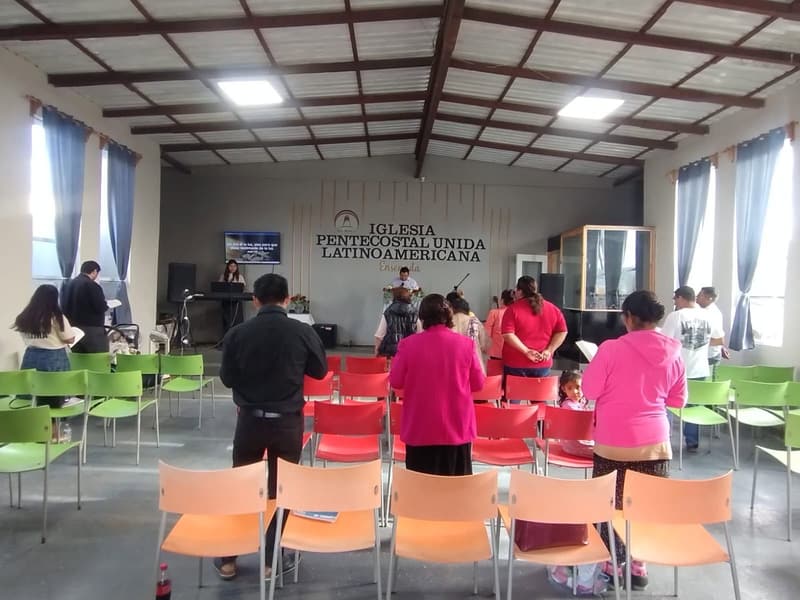 Personas adorando de pie en el salón de la iglesia de Ensenada, vistas de espaldas, frente al escenario y el nombre de la iglesia.