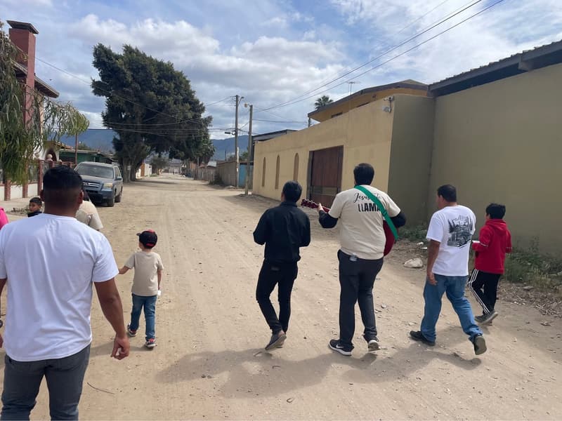 Miembros y niños caminando por una calle de terracería de un barrio, uno de ellos con una guitarra, vistos de espaldas.