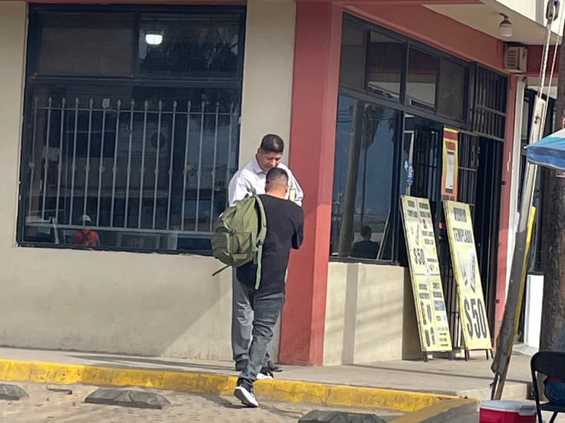 Un miembro de la iglesia platica con un hombre que lleva una mochila, afuera de una tienda, visto de espaldas.