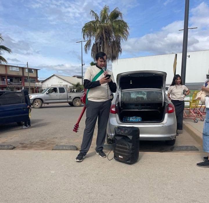 Un joven con una guitarra a la espalda, de pie junto a la cajuela abierta de un coche y una bocina en un estacionamiento, con una mujer cerca.