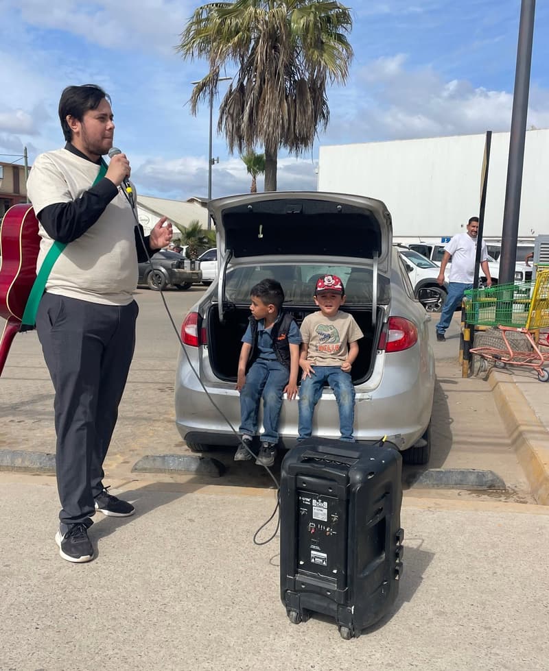 Un joven predica con un micrófono en un estacionamiento, con una guitarra a la espalda, mientras dos niños están sentados en la cajuela abierta de un coche junto a una bocina.