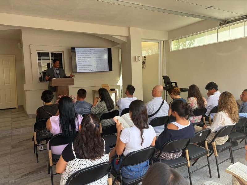 Un servicio en IPUL Tijuana: un hombre predica en el púlpito, con versículos de la Biblia en inglés y español en la pantalla detrás de él, mientras la congregación escucha, algunos con la Biblia abierta.